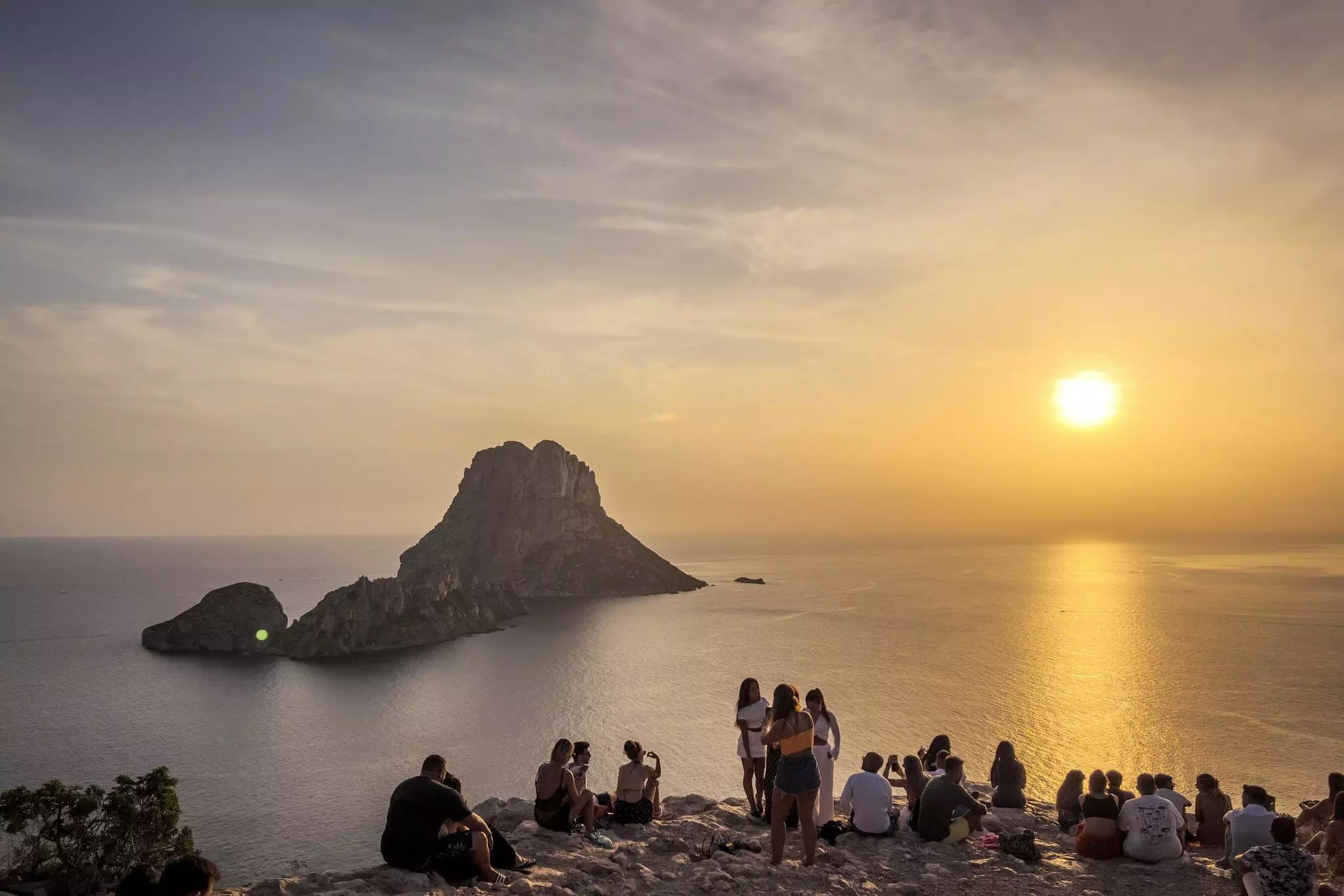 People look out at a rocky island and the sea at sunset.