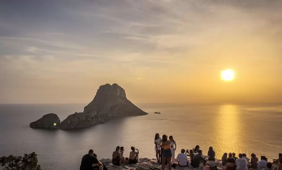 People look out at a rocky island and the sea at sunset.