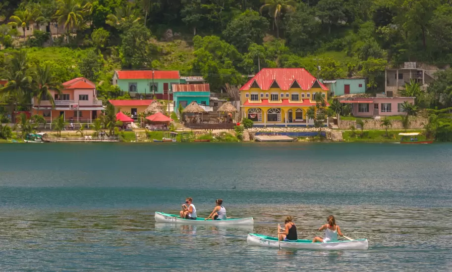 Two canoes paddled on a lake in Guatemala past brightly painted red-roofed houses and palms on a green hillside