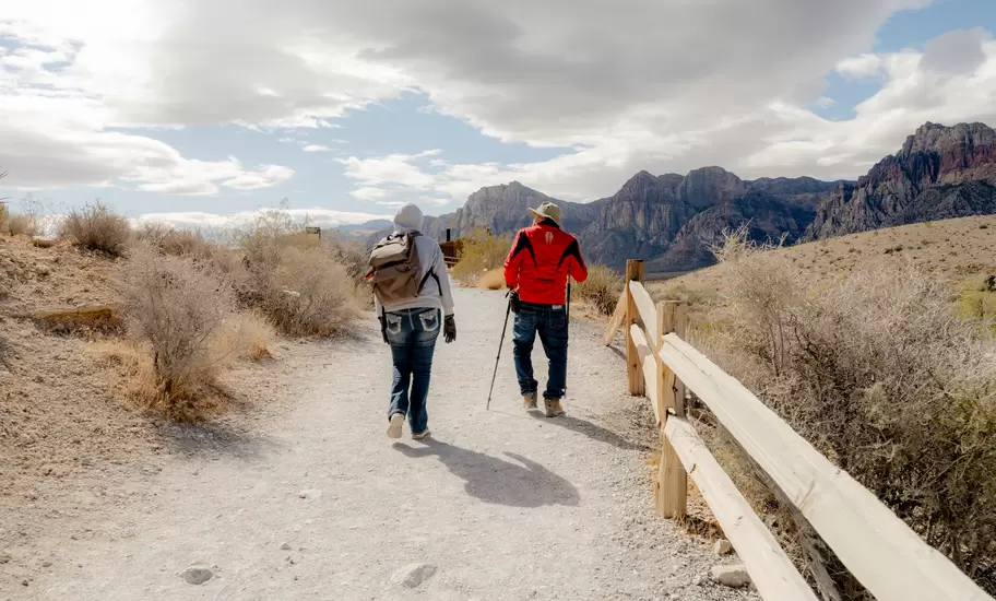 Two hikers walk a gravel trail beside a wooden fence toward rugged red-and-grey desert mountains in Nevada