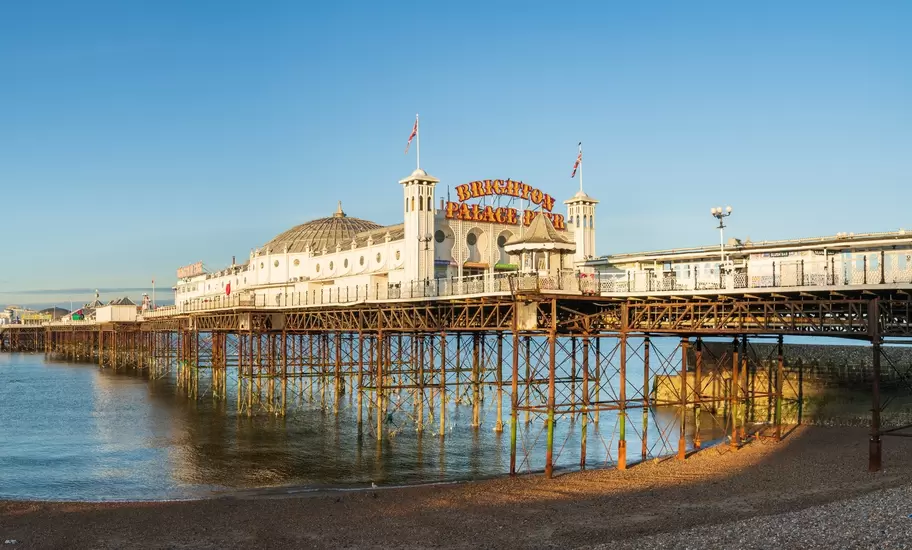 Brighton Palace Pier with its domed pavilion, flags and lit sign on iron legs, seen from the shingle beach