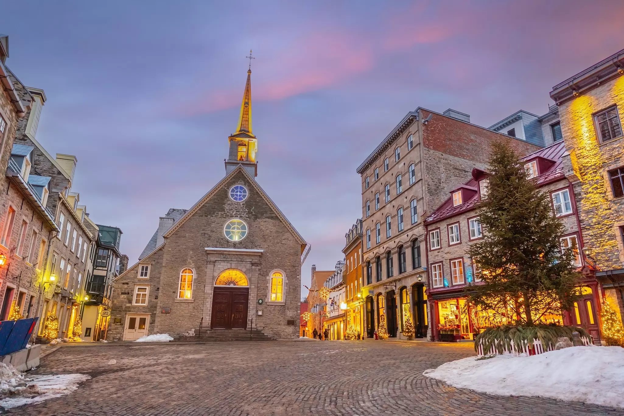 A stone church is the centerpiece of a city square surrounded by other historic buildings, pictured at dusk, Québec City, Canada
