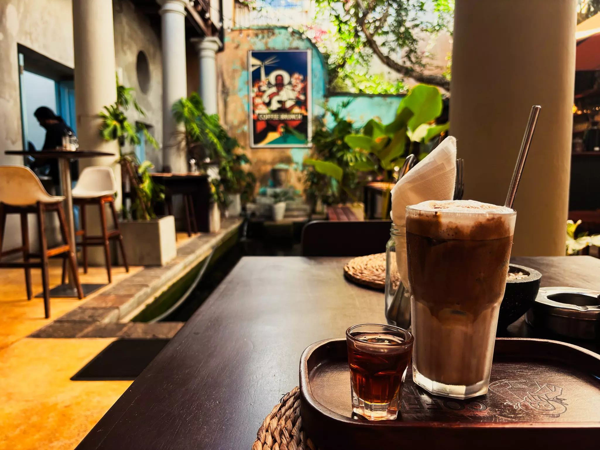 A glass of iced coffee on a wooden table with vintage posters in the background.