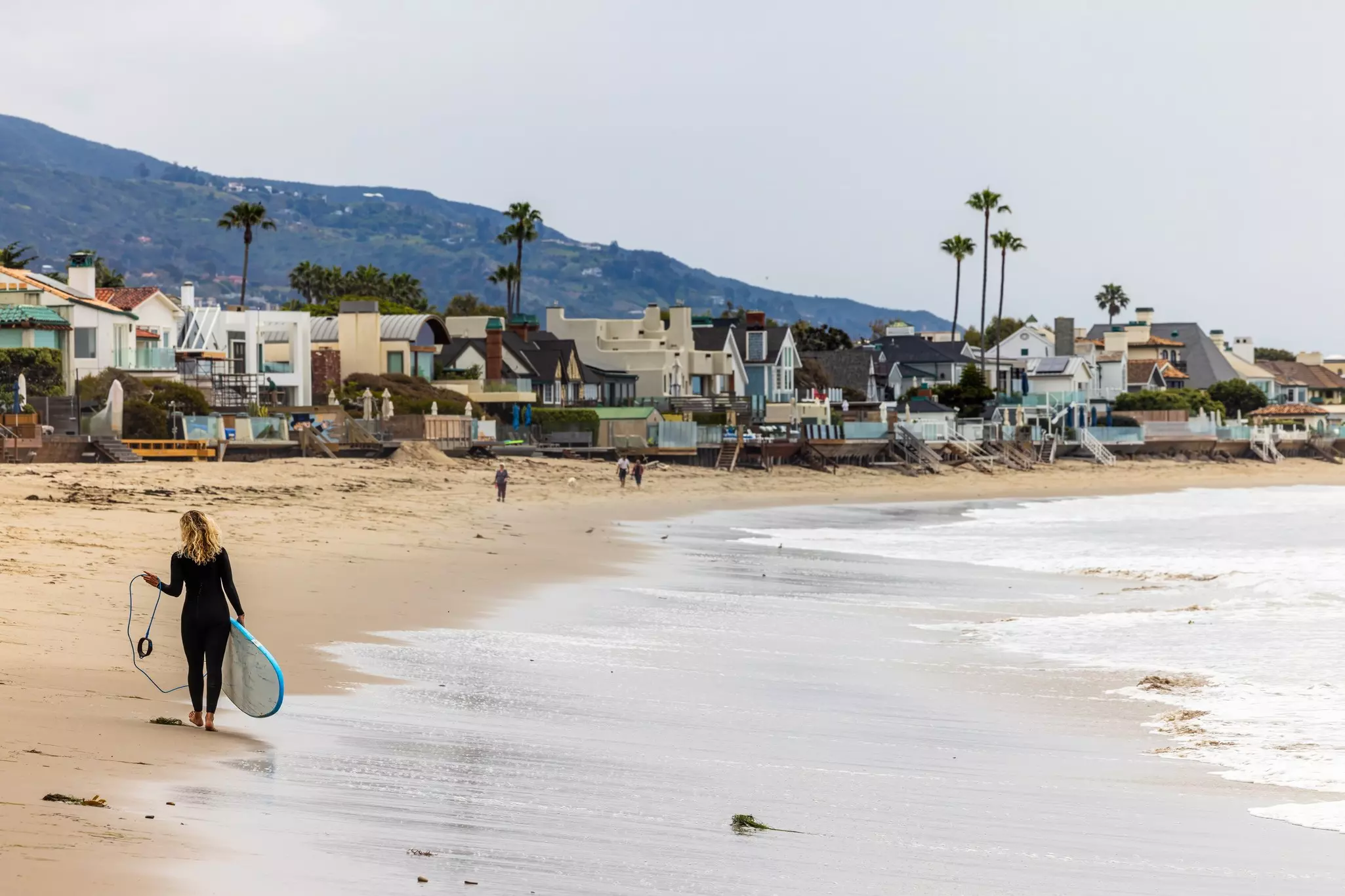 A surfer in a wet suit walks along a sandy beach carrying a surfboard.