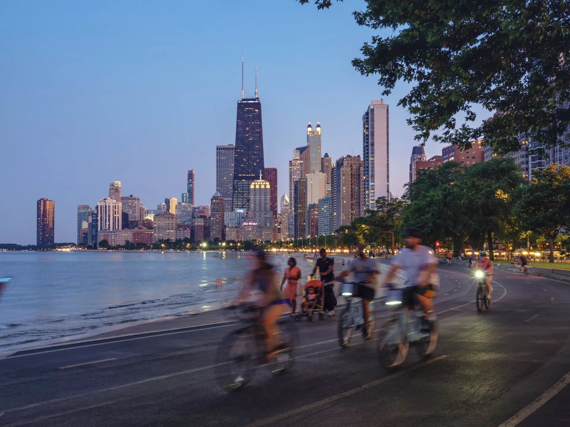 People on bicycles following a trail beside a lake, with skyscrapers in the background