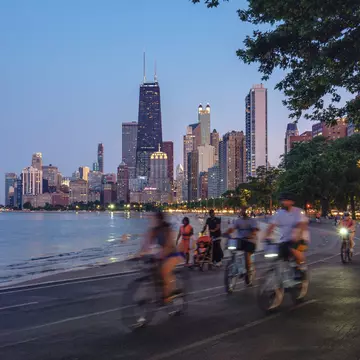 People on bicycles following a trail beside a lake, with skyscrapers in the background