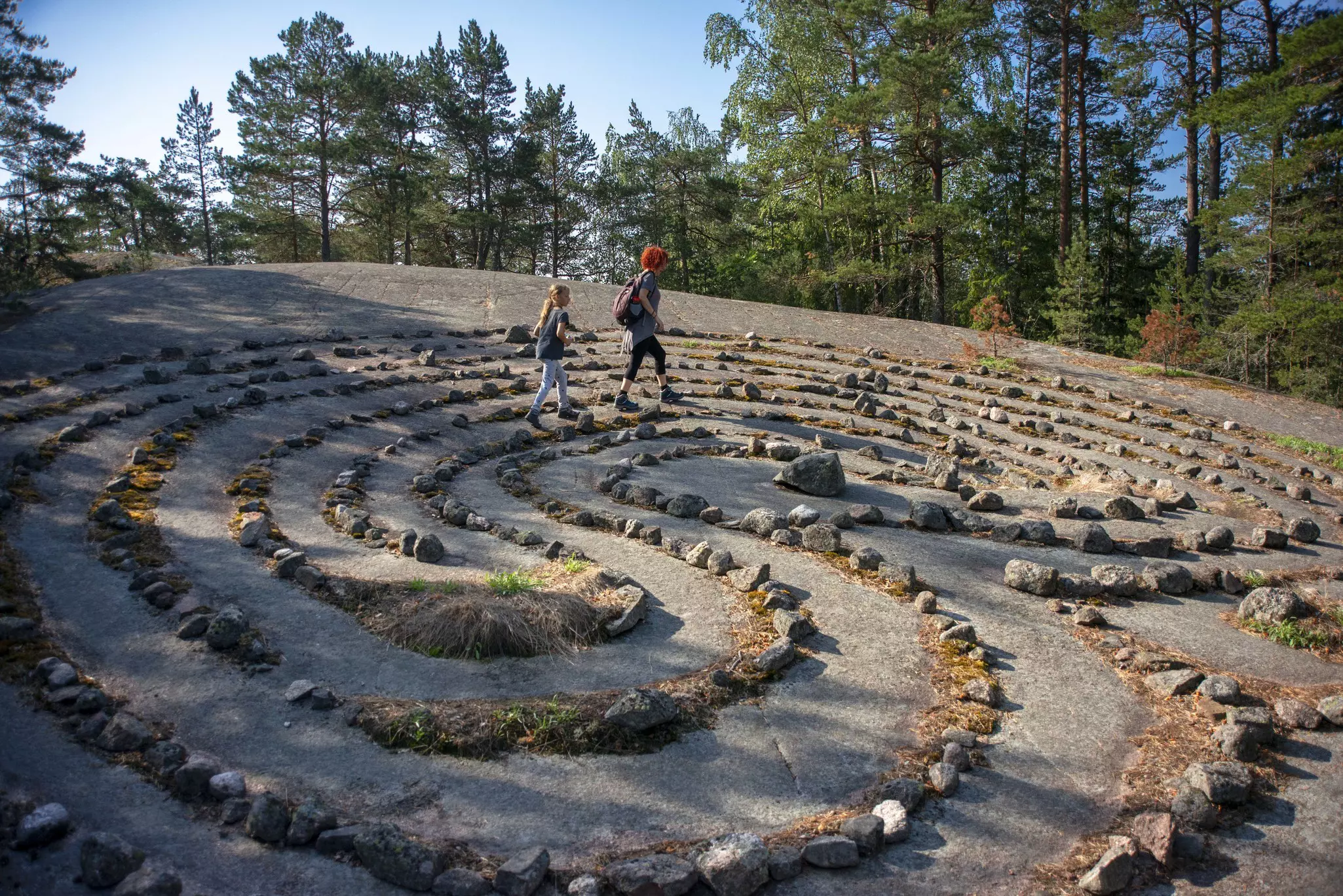 A woman a young girl walk a large stone labyrinth on a sunny day.