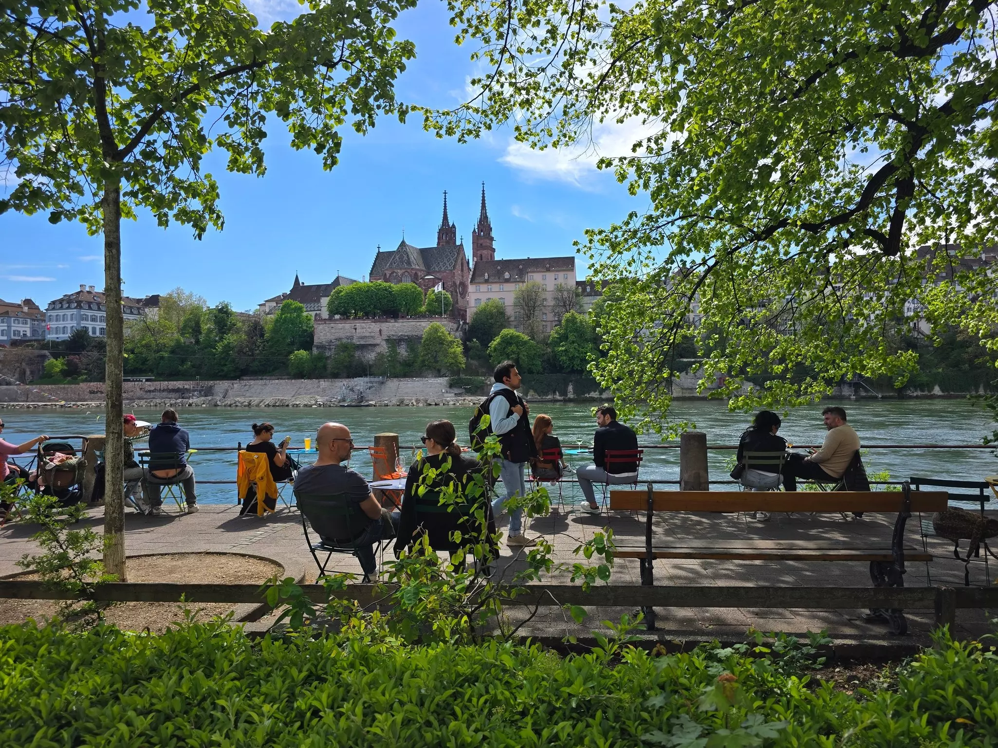 People sitting on benches and chairs on a riverfront with a view of a city on the other side