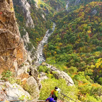 Two hikers with backpacks on a steep metal stairway beside granite cliffs, looking down a forested valley in autumn colours.