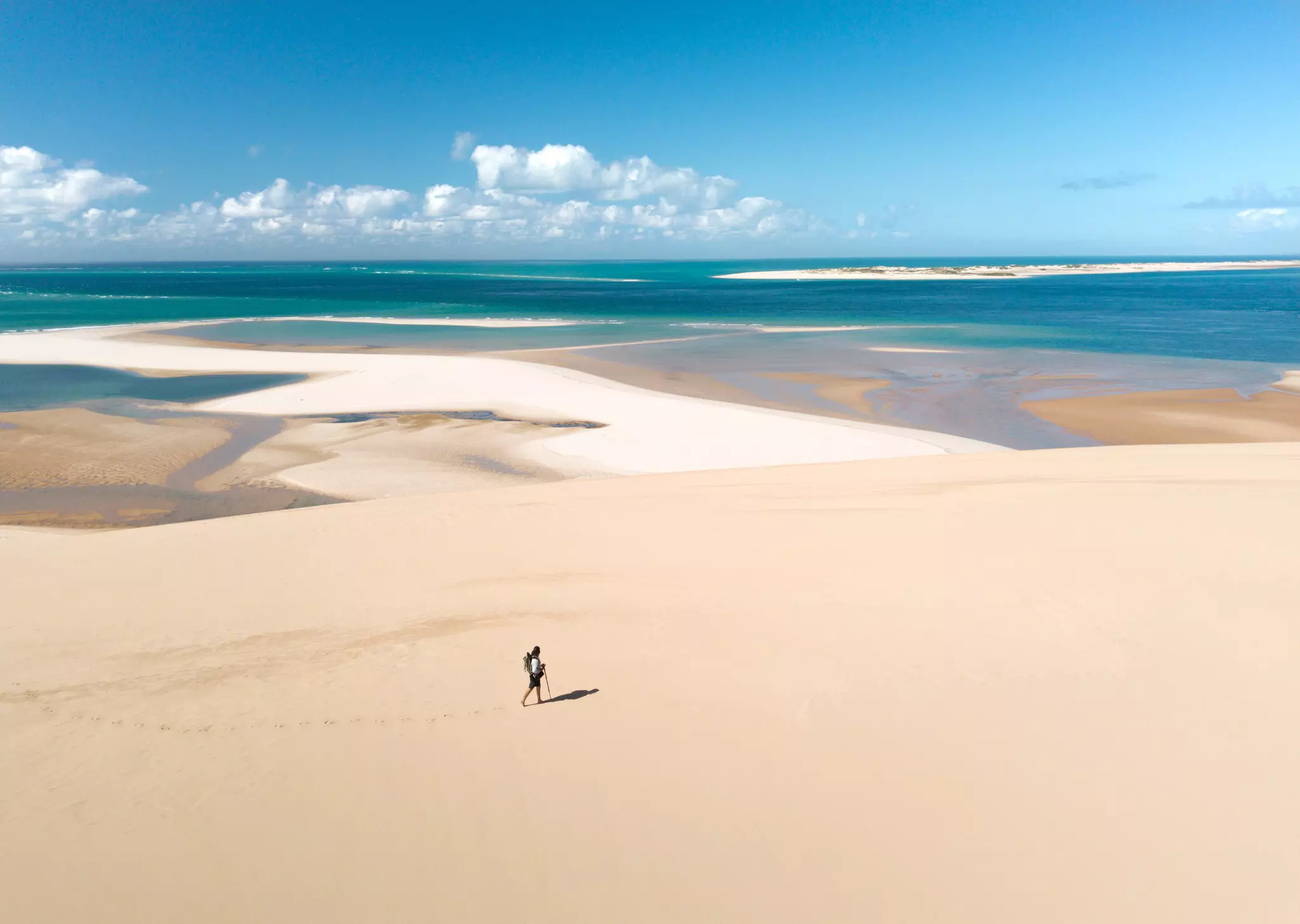 A hiker walks on a large expanse of sand with turquoise ocean in the background