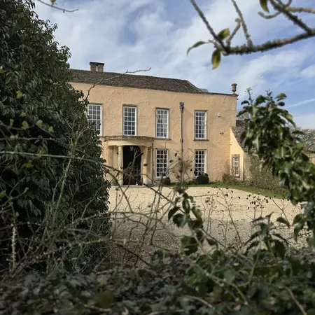 A two-story tan stone house with a rounded portico over the central door, a gravel area in front and green shrubbery in the foreground.