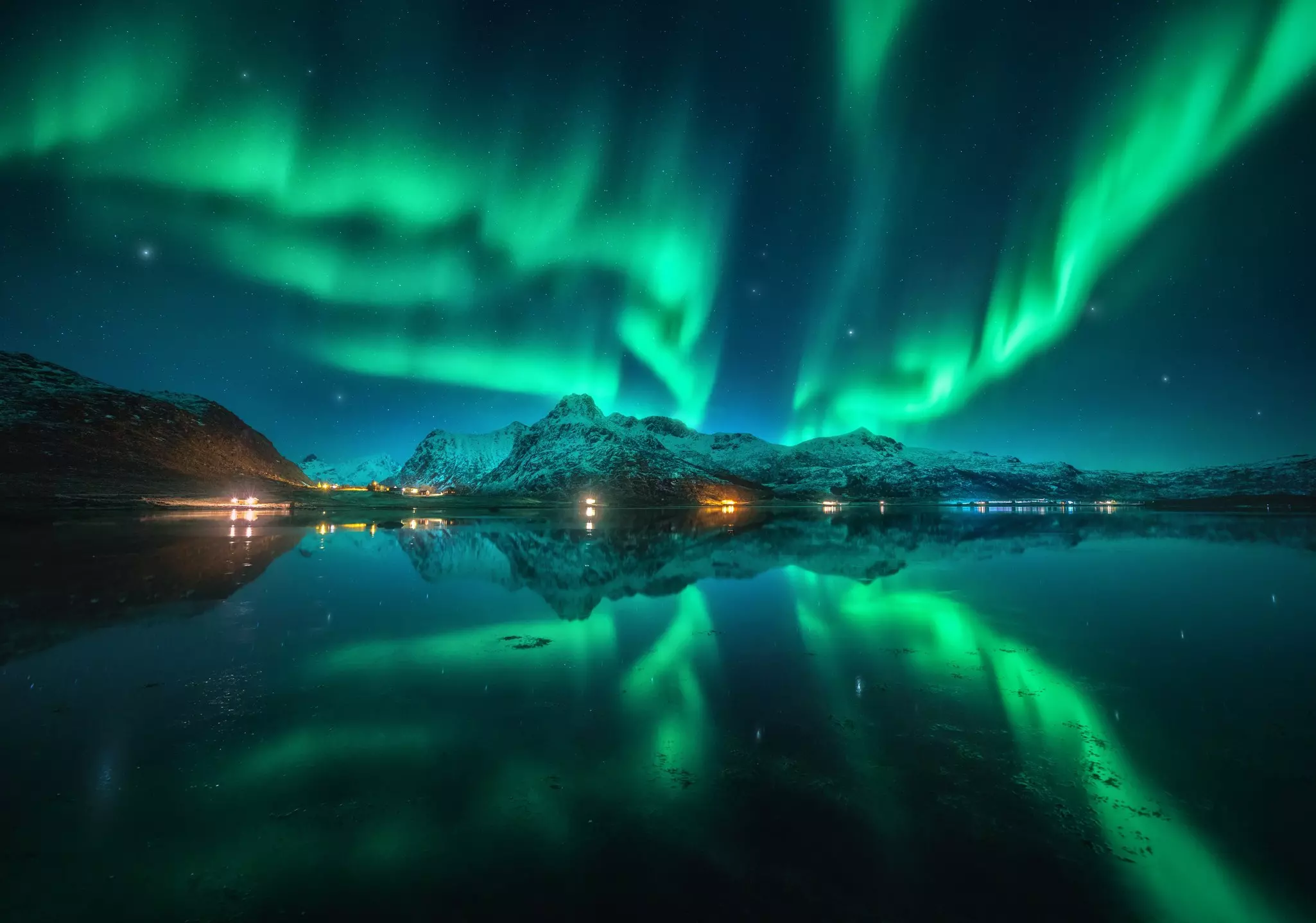 Green lights dance in a night sky above snow-covered mountains and reflected in water below.