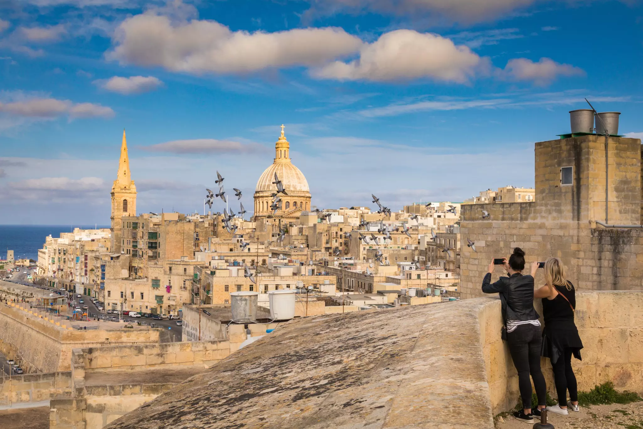 Two women stand at a city viewpoint taking photographs over the dome of a church and surrounding buildings as a flock of pigeons fly by
