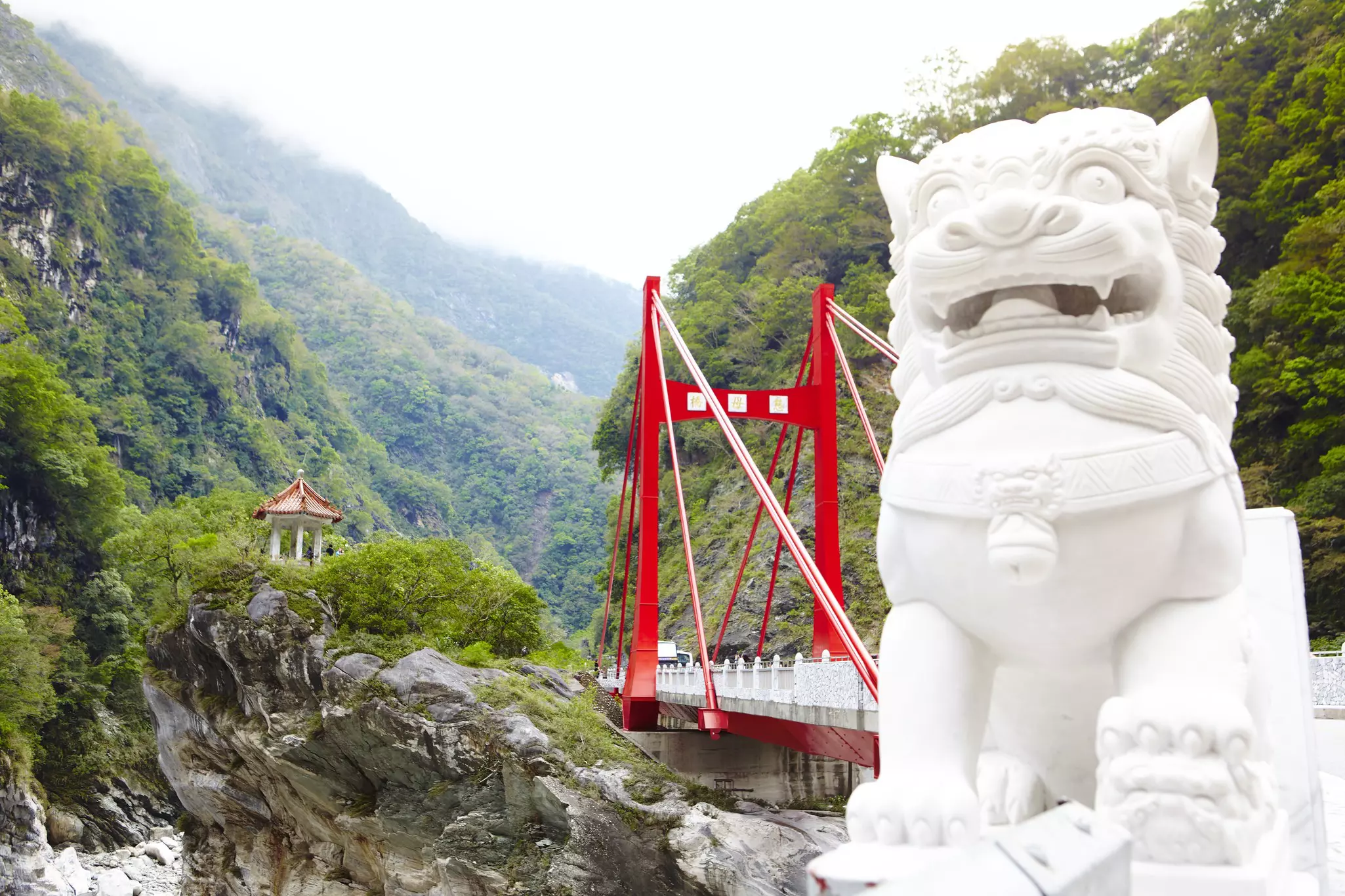 A white stone lion at the edge of a red bridge crossing part of a high-sided gorge