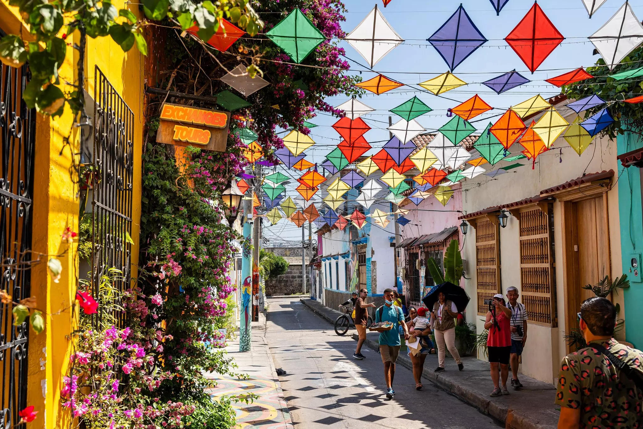 People walk down an alley with colorful paper kits strung above head height and bougainvillea growing by brightly painted walls
