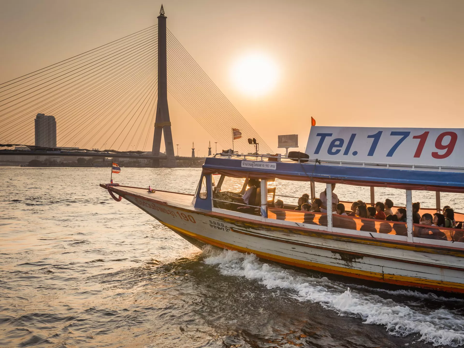 Passengers ride a ferry boat on a river in a city at sunset. A bridge is seen in the distance.