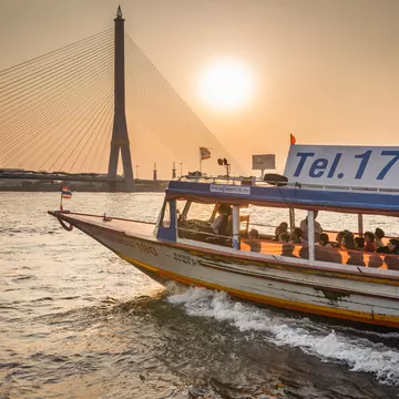 Passengers ride a ferry boat on a river in a city at sunset. A bridge is seen in the distance.
