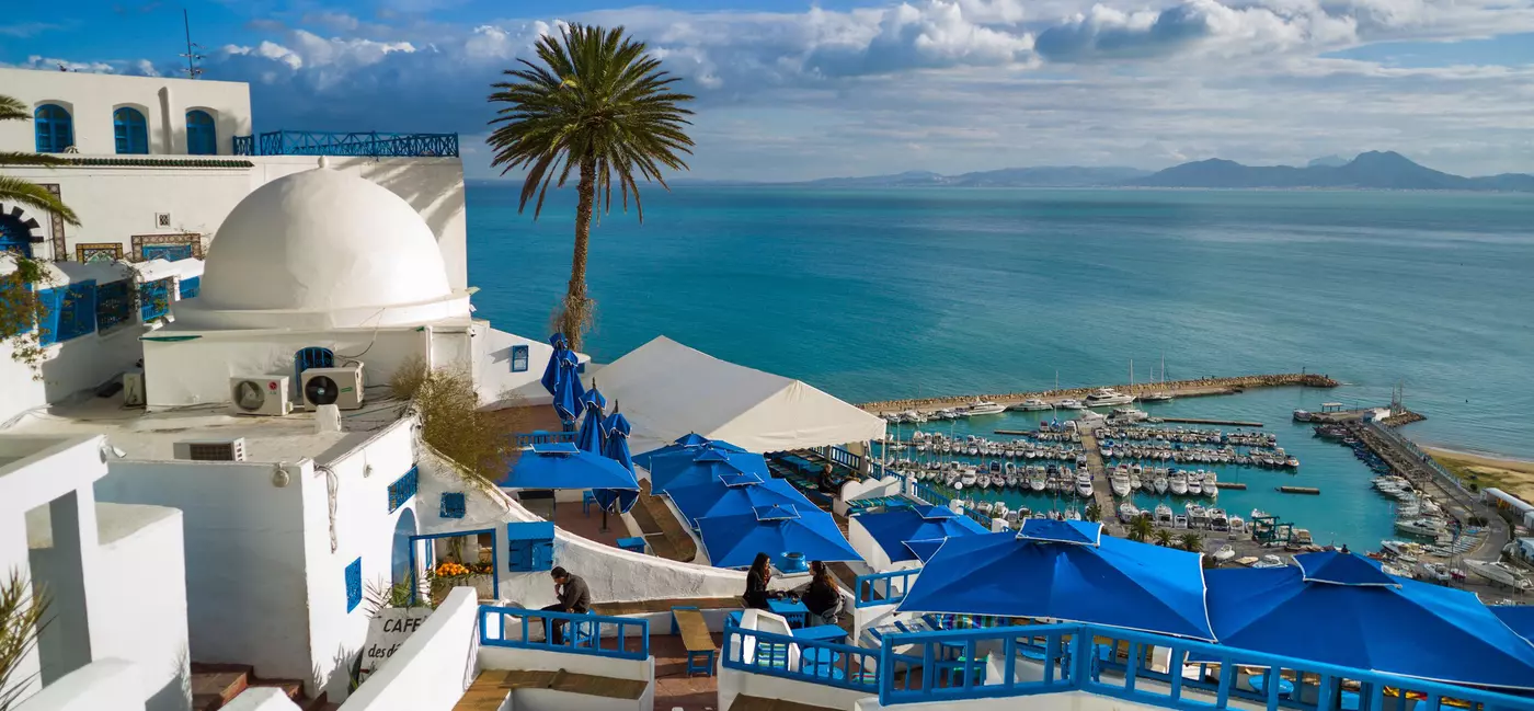 A cafe overlooking a marina and lined with blue tables and wide blue parasols.