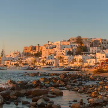 White houses stacked up a hill to a castle at the top, sailboat masts in the harbour and rocks in the foreground at sunset.