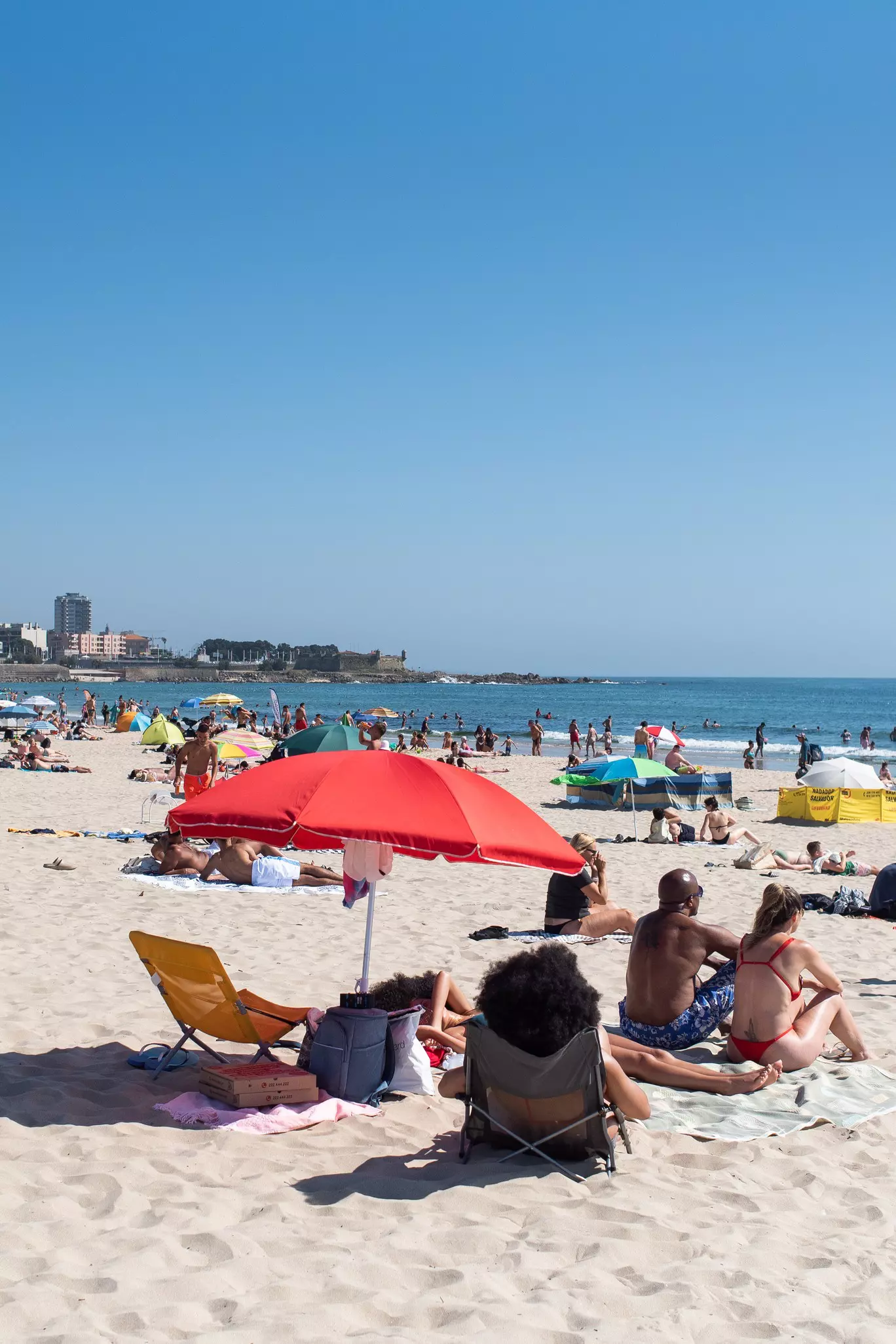 People relax under a red umbrella that is standing on the sand on a busy city beach on a sunny day.