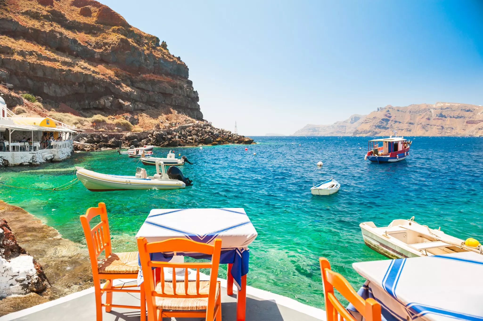 Orange chairs at a table with a white tablecloth with blue stripes is outdoors on a patio by the sea, where boats are in the harbor.