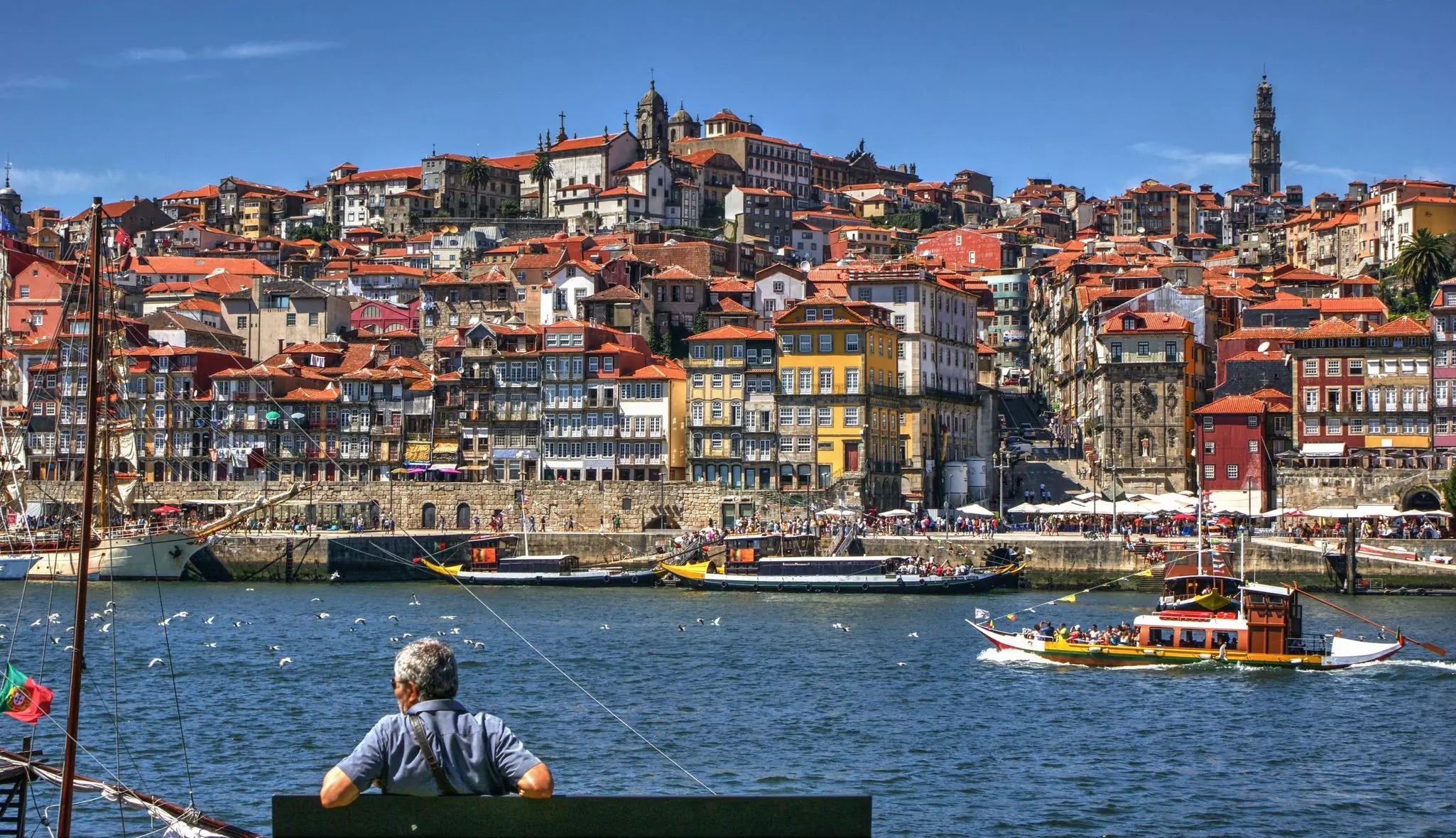 A person on a bench by a river with boats in the water; on the other shore, a city's historic buildings are built on a hillside.