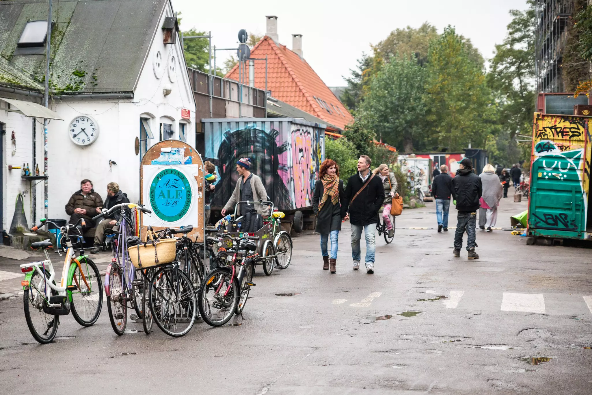 Street scene at the autonomous Freetown Christiana, in Christianshavn neighbourhood. Some tourists are visible in the image, walking around or sitting by a building having a conversation. The Freetown being car free, bicycles can also be see in the image.