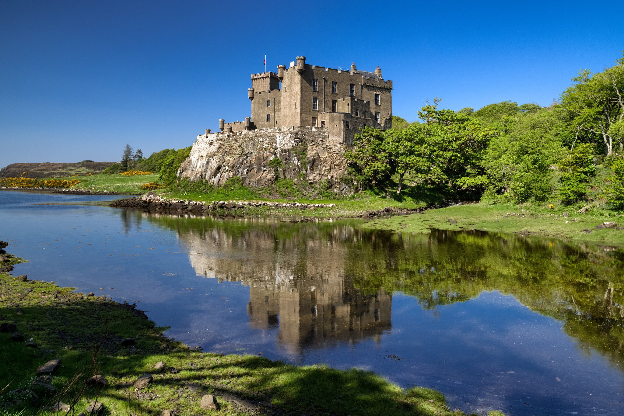 A grey turreted castle standing on a rock outcrop above still water that mirrors it, with gorse and oak woods along the shore.