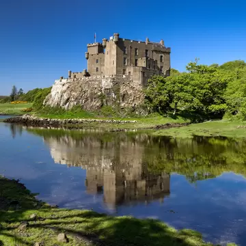 A grey turreted castle standing on a rock outcrop above still water that mirrors it, with gorse and oak woods along the shore.