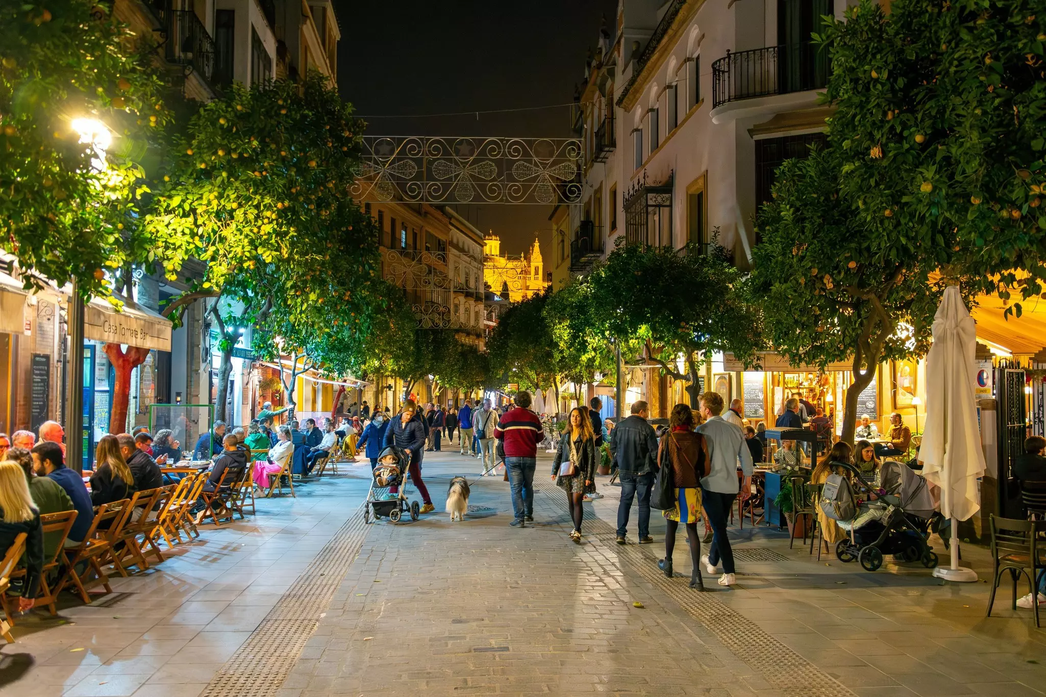 People walk and eat at cafe tables on a pedestrian street at night.