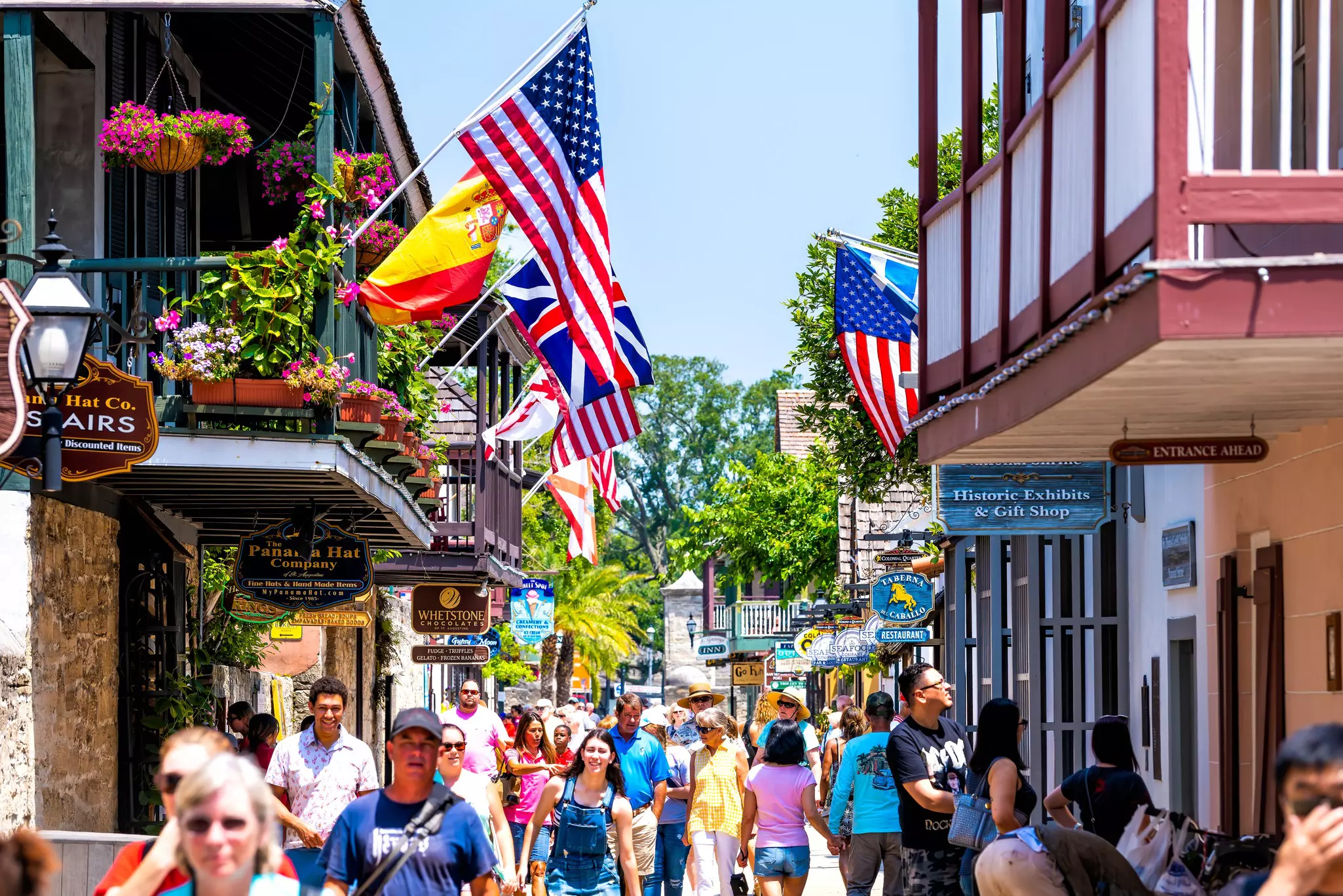 People walk past stores and under a display of flags on a pedestrianized street in a city on a sunny day.