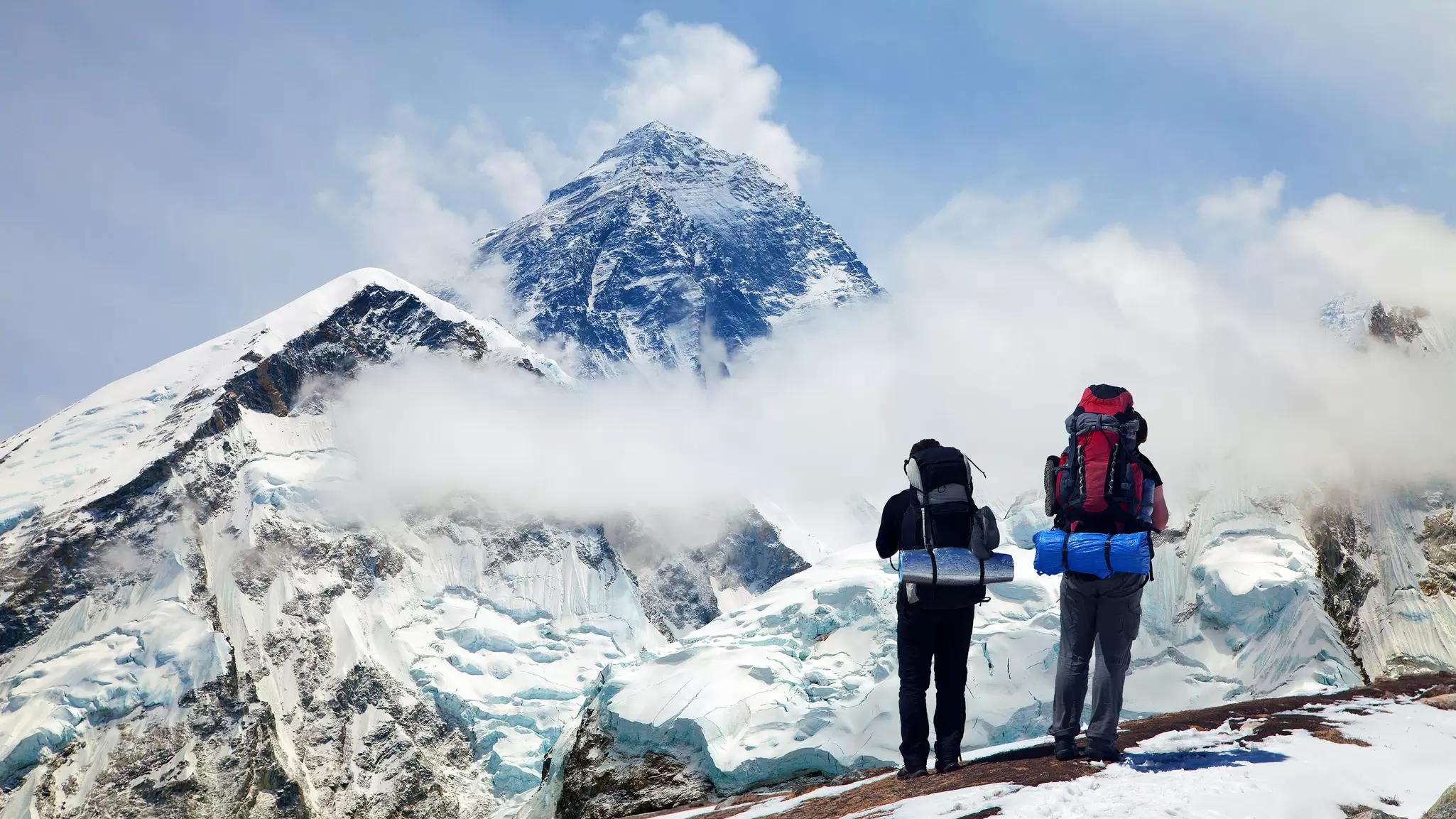 Two hikers with backpacks stand on a snowy overlook facing a tall mountain.