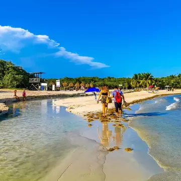 A couple walks along wet sand between a clear freshwater channel and the sea, with palapa umbrellas and palms behind