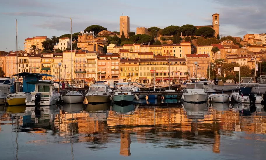 Boats moored in a harbor at golden hour before a hillside old town topped by a stone tower and clock tower