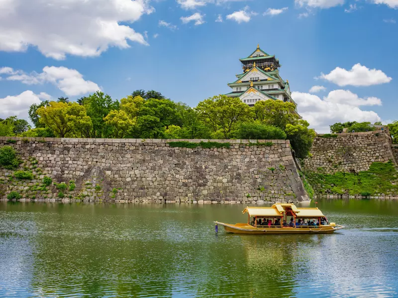 A traditional Japanese structure rises above green trees that are planted above a stone wall. The stone wall abuts a river on which a boat carries tourists.