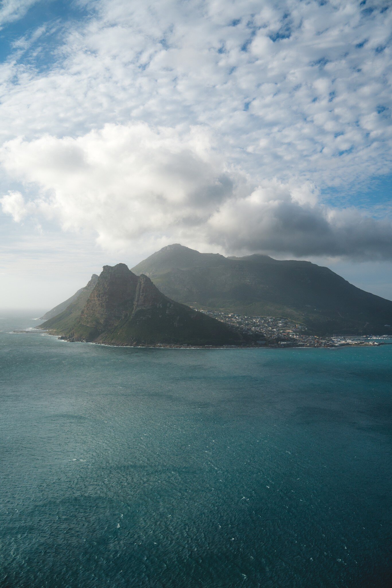 A rocky headland and mountain above the small town of Hout Bay, Cape Town, across blue-green ocean under broken cloud.