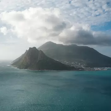 A rocky headland and mountain above the small town of Hout Bay, Cape Town, across blue-green ocean under broken cloud.