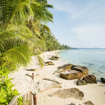 Wooden steps down to a white-sand beach lined with coconut palms and boulders, with two children near the clear sea.