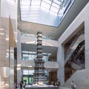 Tall carved stone pagoda beneath a vast skylight, visitors gathered at its base beside escalators
