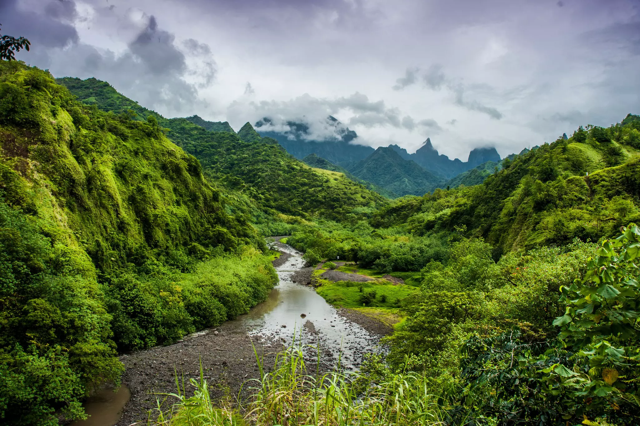 A river runs through a lush valley with clouds skipping over the top of the distant mountain peaks.