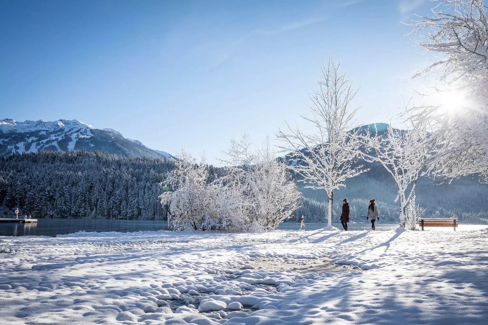 Two women walking by a lake through a snowy park with snow covered trees.