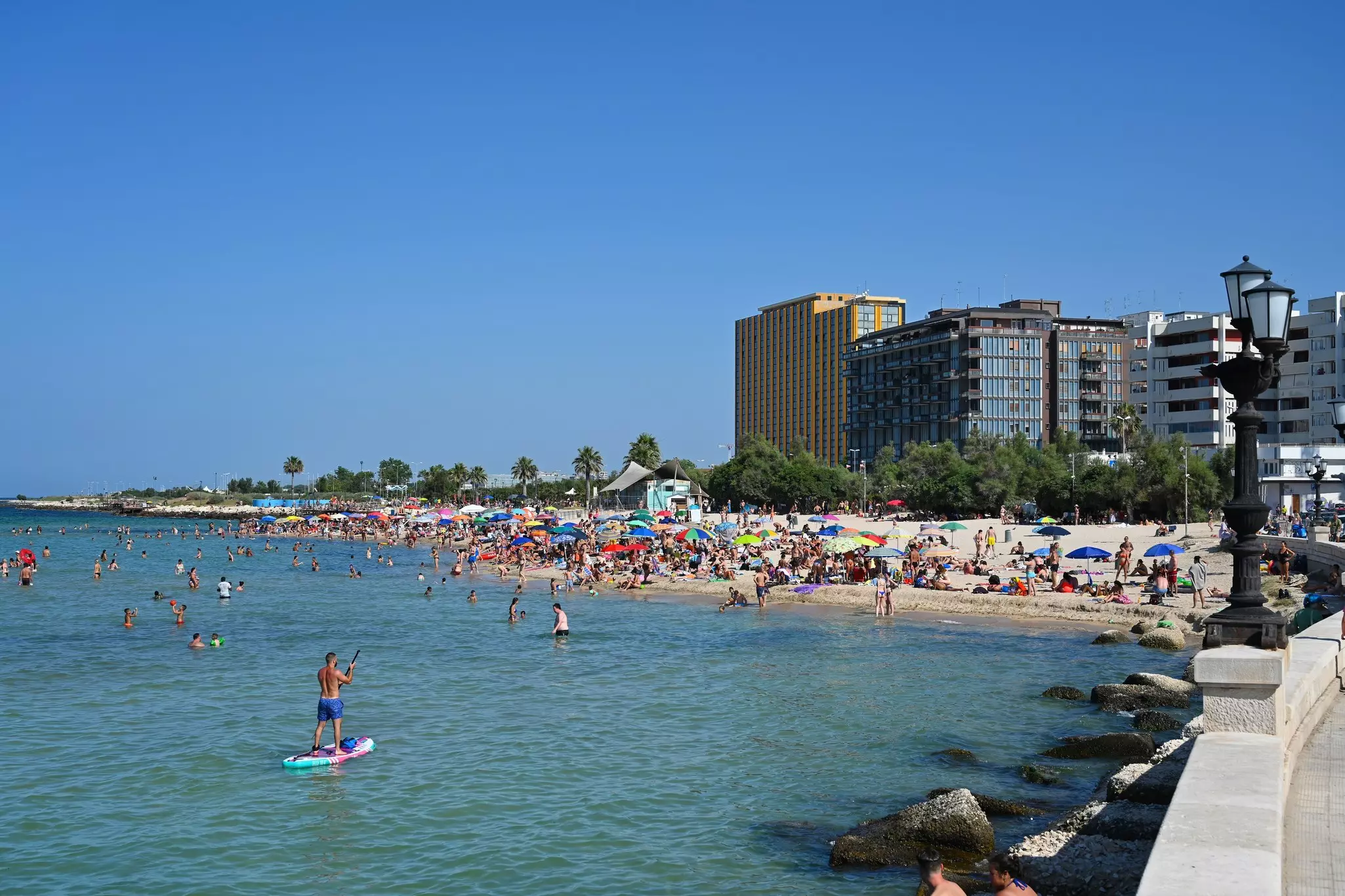 People swim and stand on paddle boards in the water off a crowded city beach.