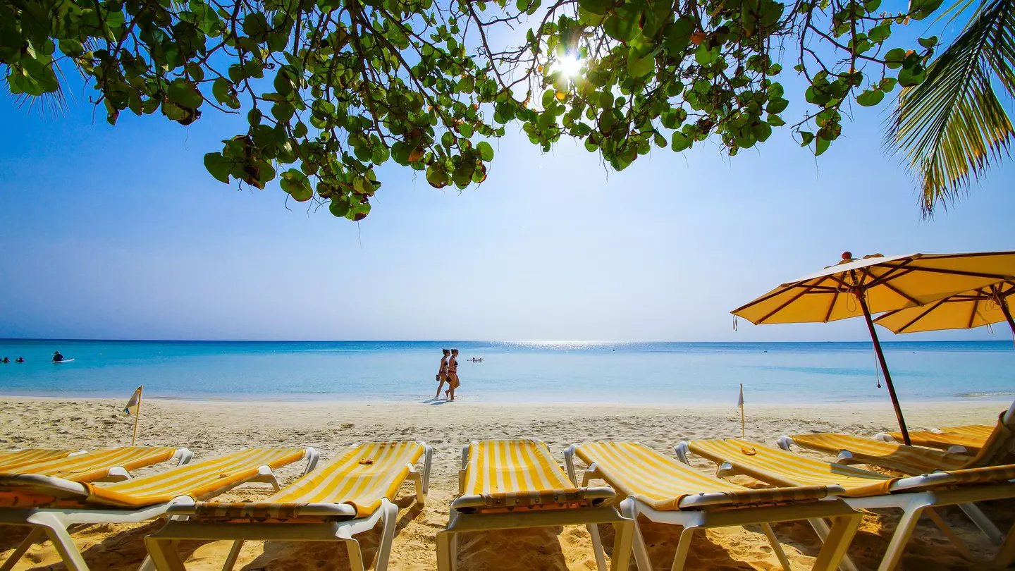 Chaise lounges with yellow-striped towels sit on a beach with blue waters. People walka long the beach in the distance.