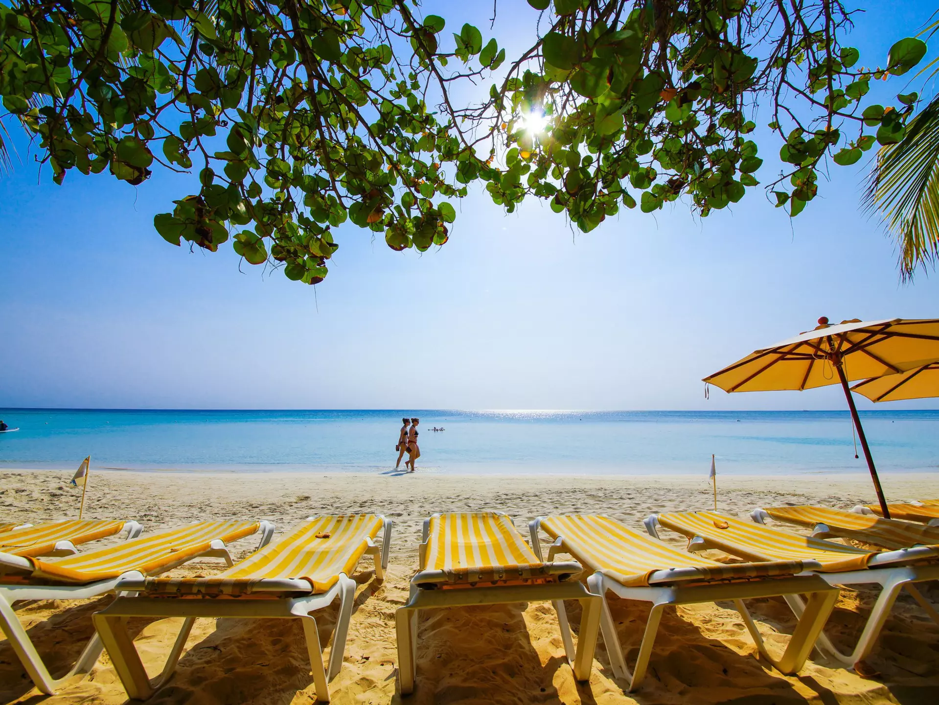 Chaise lounges with yellow-striped towels sit on a beach with blue waters. People walka long the beach in the distance.