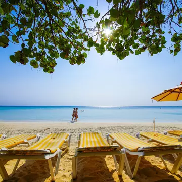 Chaise lounges with yellow-striped towels sit on a beach with blue waters. People walka long the beach in the distance.