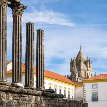 Weathered Corinthian columns in the foreground with a pointed stone cathedral tower and orange-roofed buildings, Evora.