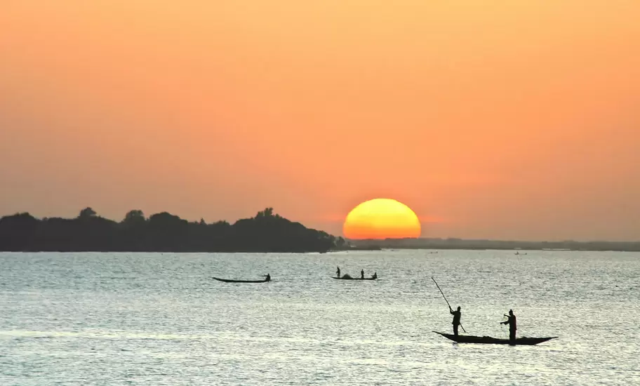 Silhouetted fishers poling wooden pirogues across a wide river in Niger as an orange sun sets behind a wooded island