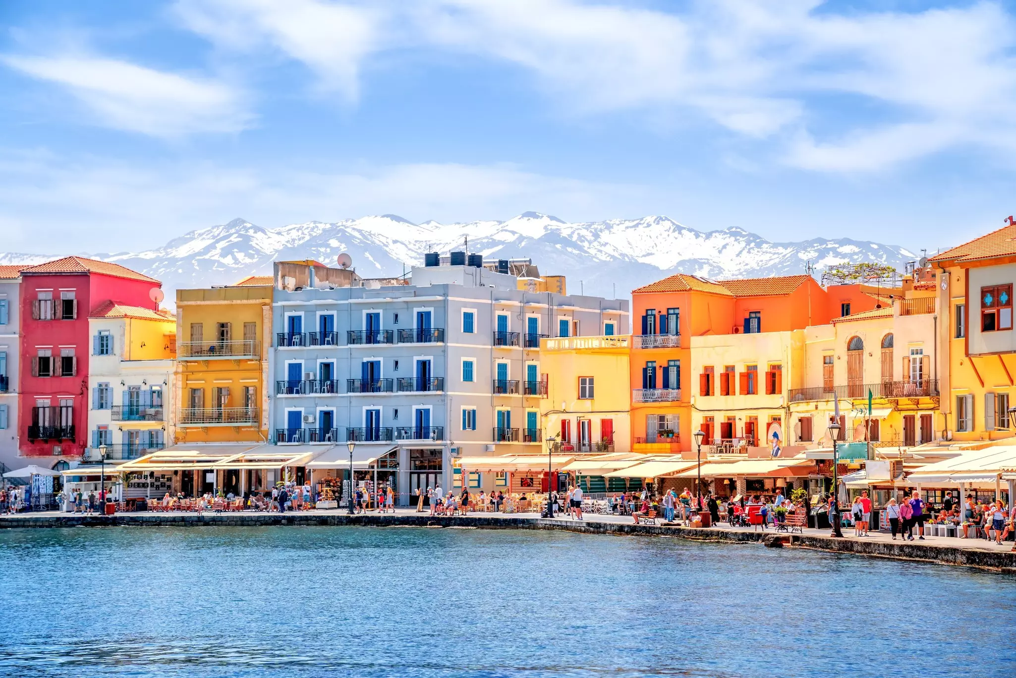 Colourful buildings and open air restaurants on a Greek waterfront with snowy mountains in the background