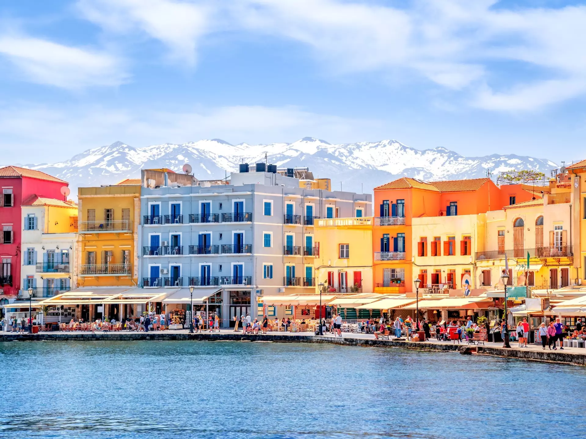 Colorful buildings and open-air restaurants on the waterfront in Hania, Crete, with snowy mountains in the background.