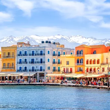 Colorful buildings and open-air restaurants on the waterfront in Hania, Crete, with snowy mountains in the background.